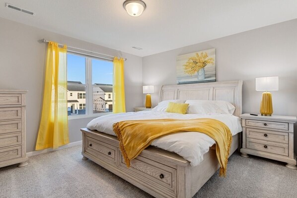 Un dormitorio con una cama grande, dos mesitas de noche, una ventana con cortinas amarillas y un cuadro en la pared.