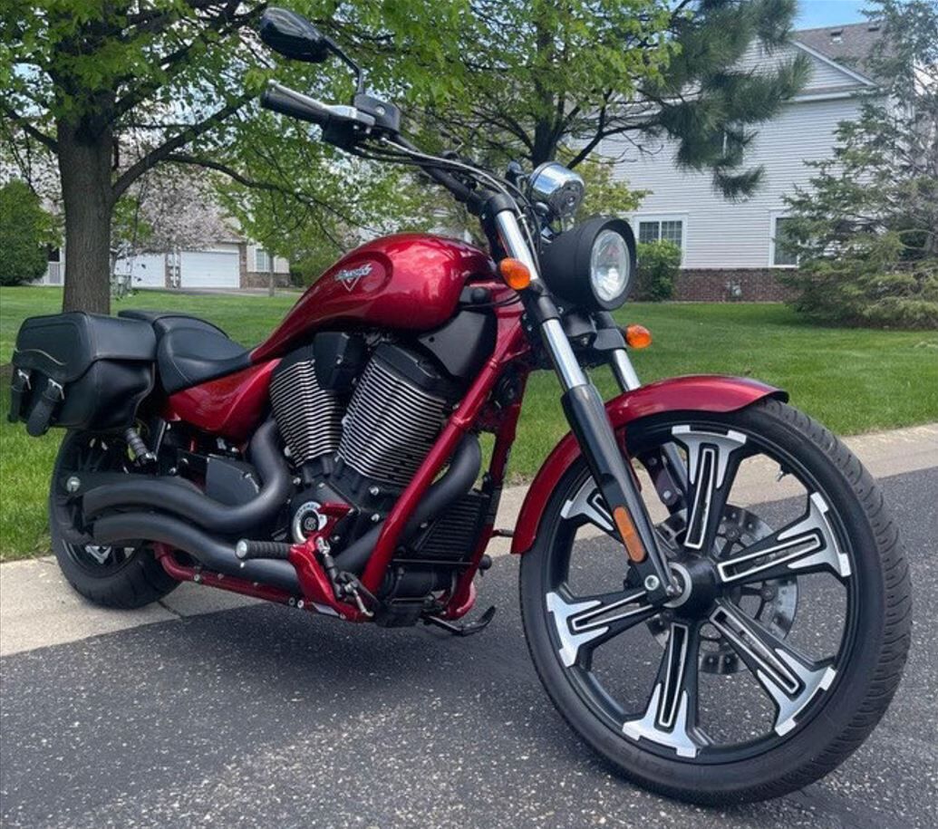 A red motorcycle parked on a paved surface with a house and trees in the background.