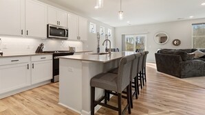 Een moderne keuken met witte kasten, een centraal eiland met barstoelen, een magnetron en een spoelbak. In de woonkamer staat een donkergrijze bank en een groot raam.