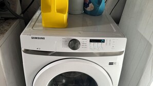 Una lavadora Samsung con puerta frontal, un panel de control con varias opciones y botellas de detergente encima.