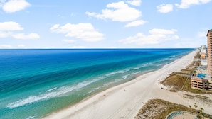 Een strand met helderblauw water, een zandstrand en een hotel met meerdere verdiepingen en balkons met uitzicht op het strand.