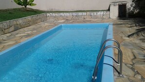 A rectangular swimming pool with a metal ladder on one side, surrounded by stone tiles and a low stone wall. - Alugo Casa Para Temporada em Búzios-rj (Lot. Portal da Ferradura Arm. dos Búzios)