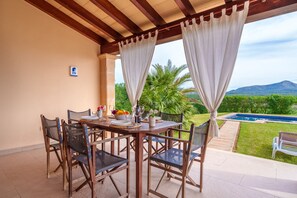 An outdoor patio with a wooden table and chairs, a pool, and a view of greenery and mountains.