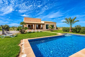 A pool with lounge chairs, a house with a tiled roof, and palm trees.