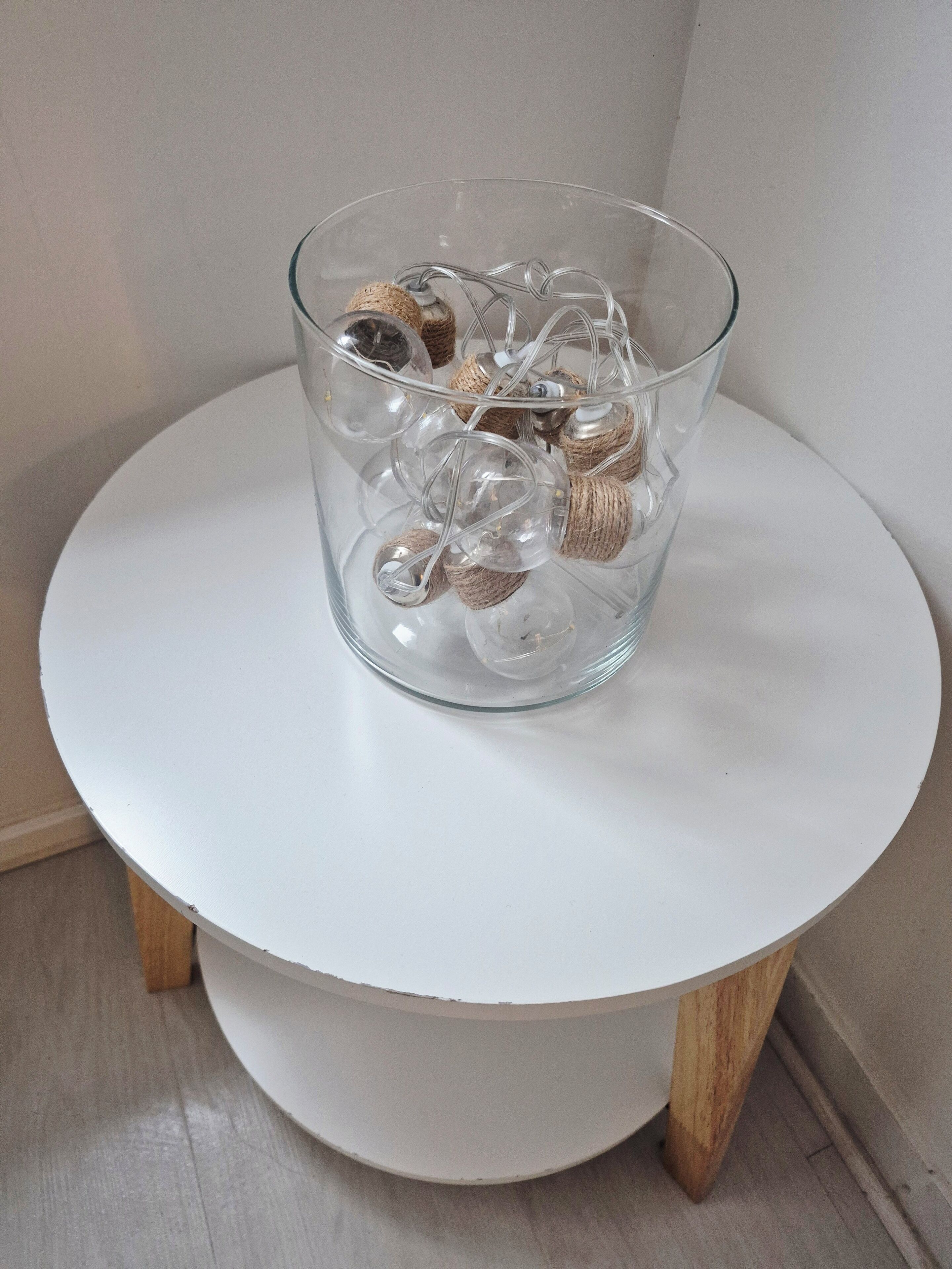 Un florero de vidrio transparente con cuerda y corchos sobre una mesa redonda blanca con patas de madera.