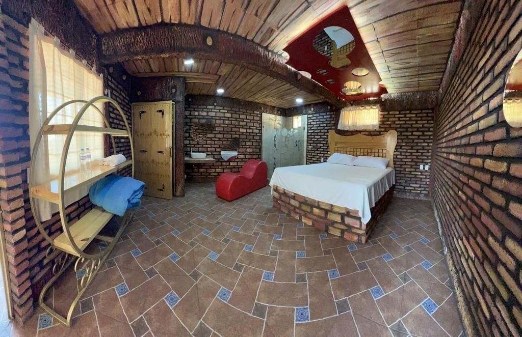 Una habitación con pared de ladrillo, una cama, una silla roja, un estante de madera y una puerta.