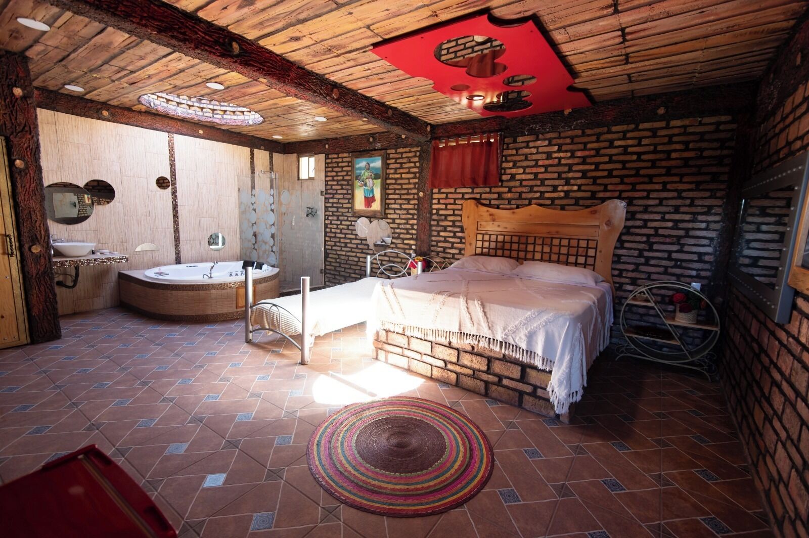 Un dormitorio rústico con pared de ladrillo, techo de madera, cama, baño pequeño con bañera y alfombra redonda.