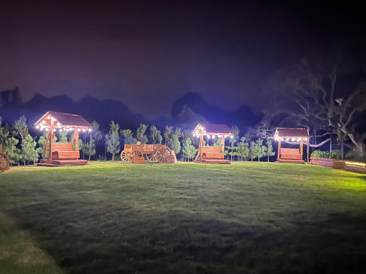 Un espacio exterior bien iluminado con estructuras de madera, posiblemente un pabellón o mirador, rodeado de árboles y un campo cubierto de césped.