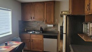 A compact kitchen with wooden cabinets, a built-in dishwasher, and a window providing natural light. - Small House in Downtown Wainwright (Wainwright)
