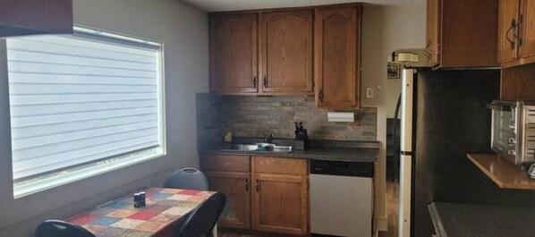 A compact kitchen with wooden cabinets, a built-in dishwasher, and a window providing natural light. - Small House in Downtown Wainwright (Wainwright)