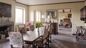 Ein Esszimmer mit Holztisch und floral gepolsterten Stühlen, einem Kamin und Blick in einen Wohnbereich mit Sofa und Lampe.
