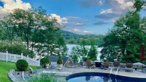 Una piscina con un solárium circundante, una cerca blanca y vistas a un lago y colinas al fondo.