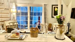 Un comedor con una mesa dispuesta para dos, un cuenco de madera, un florero con flores y una ventana con vistas a árboles.