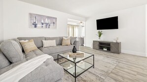 A modern living room with a sectional sofa, a glass coffee table, a flat-screen TV, and a painting of sailboats. - Relaxing Three Bedroom in PGA National (Palm Beach Gardens)