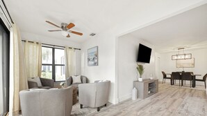 A modern living room with a grey sofa, a coffee table, and a dining area with a table and chairs. There is a ceiling fan, a flat-screen TV, and a painting on the wall. - Relaxing Three Bedroom in PGA National (Palm Beach Gardens)