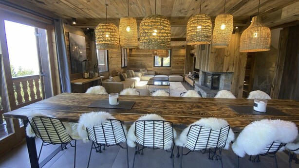 Amplia sala de estar con una gran mesa de madera, seis sillas con mantas de piel blanca y una chimenea de piedra.