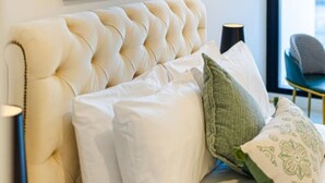 Un dormitorio con cabecera capitonada, cama con ropa blanca, almohada estampada verde y un cuadro en la pared.