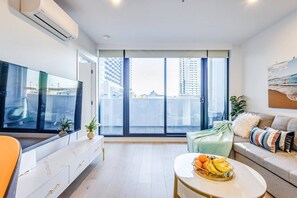 Una sala de estar moderna con un televisor grande, un sofá, una mesa de centro con fruta y un balcón con vistas a la ciudad.