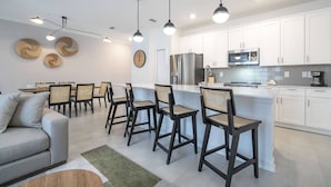 A modern kitchen with white cabinets, a stainless steel refrigerator, and a microwave. There are bar stools and a dining area with a sofa and chairs.