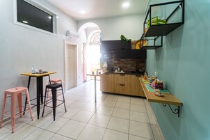Una cocina con encimera de madera, un lavamanos y una ventana sobre el lavamanos. Hay dos taburetes rosa y una mesa con cubierta amarilla.