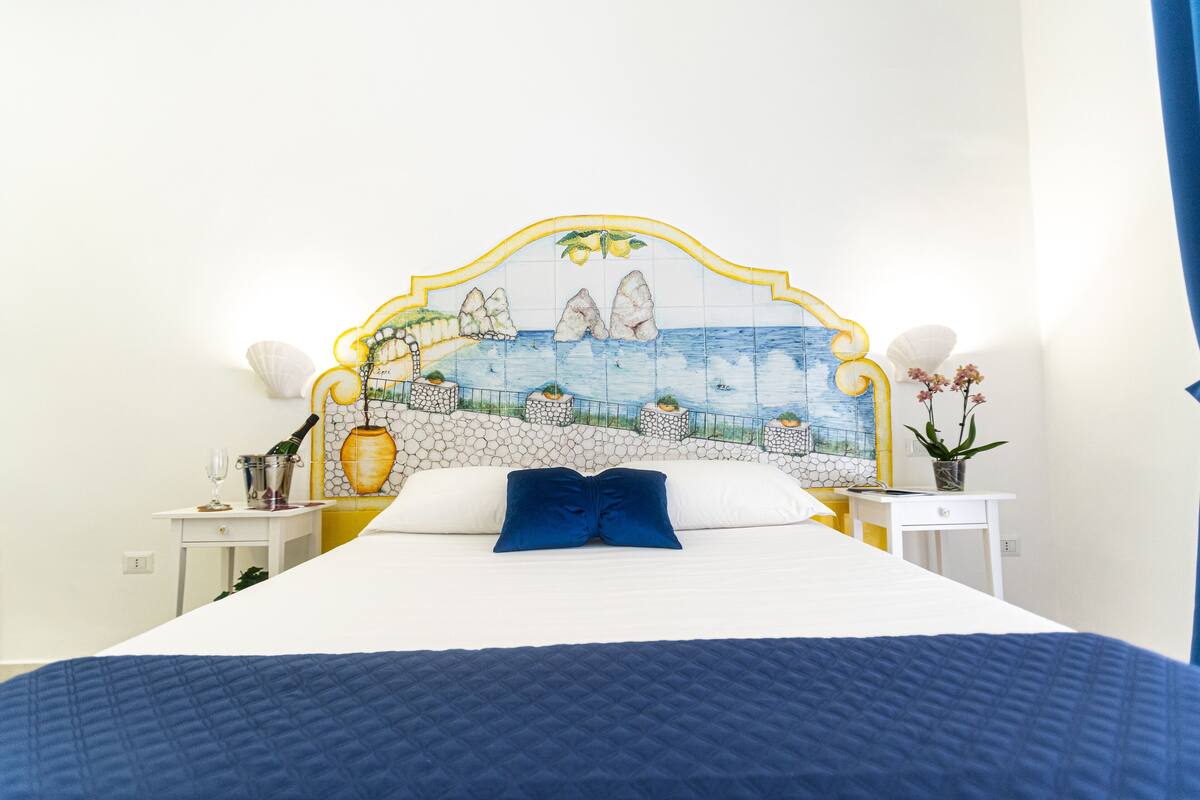 A bed with a blue quilt, a white headboard with a mosaic, and a nightstand with a vase of flowers.