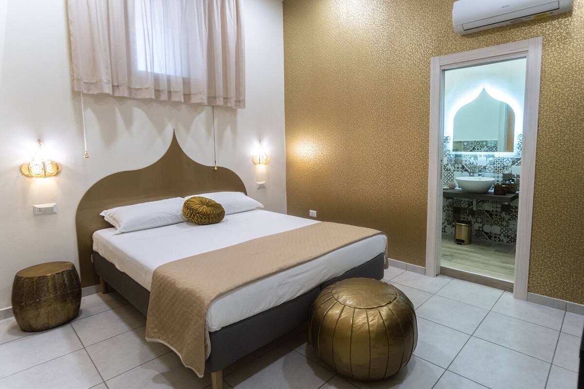 A neatly arranged bedroom with a bed, bedside lamps, a round stool, and a view of the bathroom.