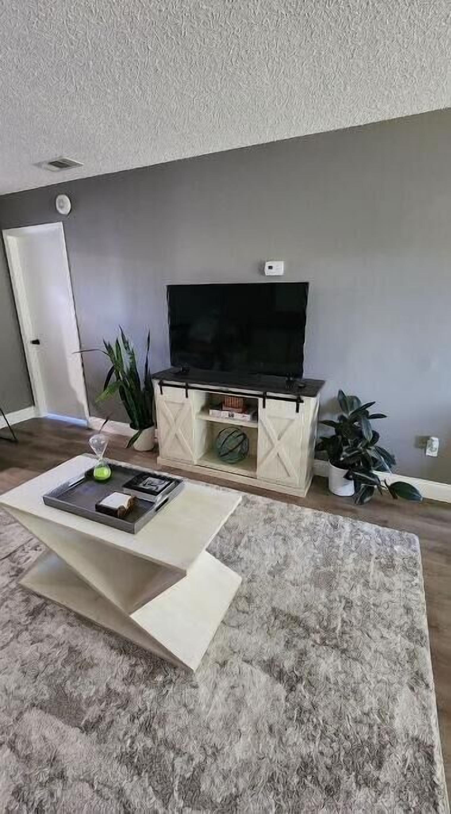 Sala de estar com televisão de tela plana em um armário branco, uma mesa de centro com revistas e duas plantas em vasos.