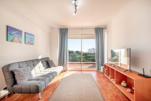 Una sala de estar moderna con un sofá gris, un televisor sobre un mueble de madera y un balcón con vista a la ciudad.