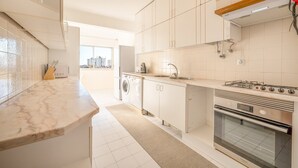 A modern kitchen with white cabinets, a marble countertop, a gas stove, a washing machine, and a view of buildings through the window. - GuestReady - Charming stay with a terrace (Cascais)
