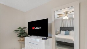 Ein Hotelzimmer mit einem Flachbildfernseher, der YouTube TV anzeigt, einer Topfpflanze, einer weißen Kommode mit Schubladen und einem großen Spiegel, der ein Bett und einen Deckenventilator reflektiert.