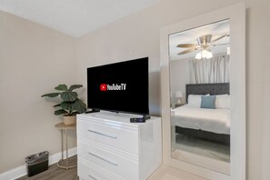 Hotelkamer met een flatscreen-tv waarop YouTube TV wordt afgespeeld, een potplant, een witte dressoir met lades en een grote spiegel die een bed en een plafondventilator reflecteert.