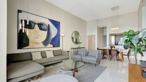 Un salon moderne avec un grand tableau abstrait, un canapé, des fauteuils, une table basse en verre et un coin repas avec des chaises et une plante.
