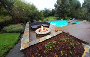 Un jardin avec un foyer en pierre, une piscine et du mobilier d’extérieur.
