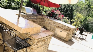 Cocina exterior con encimera de piedra, una parrilla y un lavamanos. Hay dos sillas y una mesa cerca.