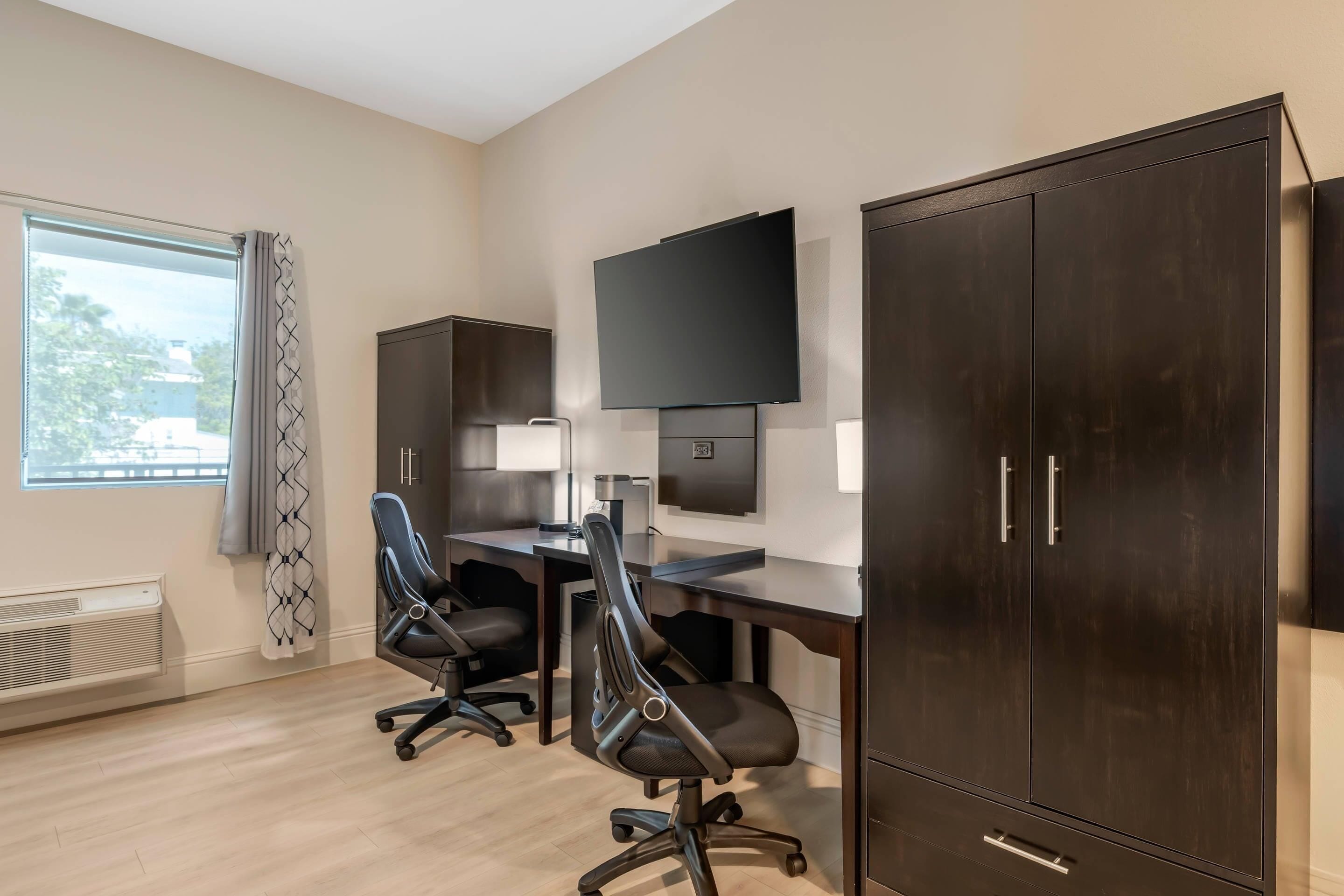 A modern office space with two black office chairs, a desk, a flat-screen TV, and dark wooden cabinets.