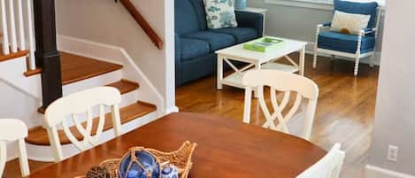 Un comedor con una mesa de madera, sillas blancas y una araña. Se observa una escalera con escalones y barandilla de madera. En el fondo, un sofá azul y una pequeña mesa con una lámpara.