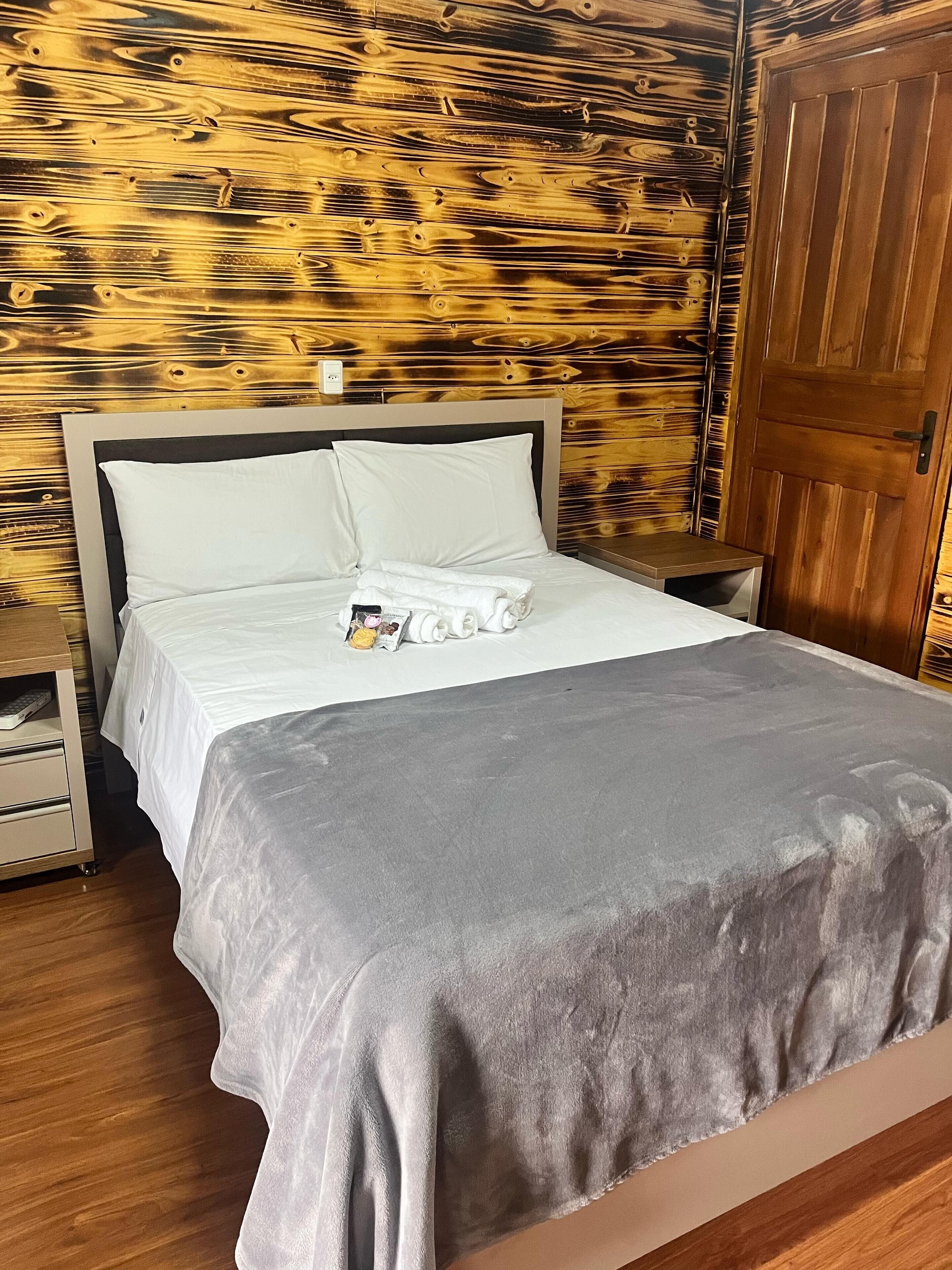 A neatly made bed with white linens and a gray blanket, a wooden nightstand with a remote control, and a wooden wall with a vertical panel design.