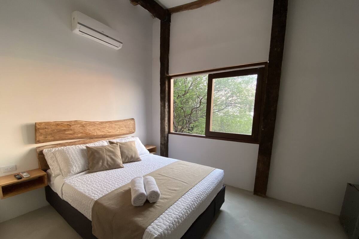 Un dormitorio con cabecera de madera, un ventanal con vistas a árboles, una mesita de noche con un teléfono y una cama con manta y almohadas color beige.