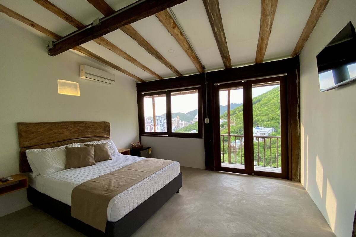 Un dormitorio con cama, vigas de madera, un ventanal y vistas a la vegetación.