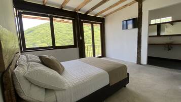 Un dormitorio con cama, ventana con vista a follaje, televisor y baño con lavabo.