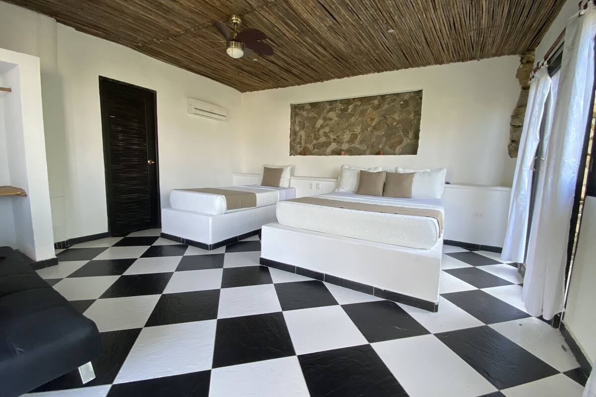 Una habitación de hotel con piso a cuadros, una cama con ropa de cama blanca, un ventilador de techo y una pared de piedra.