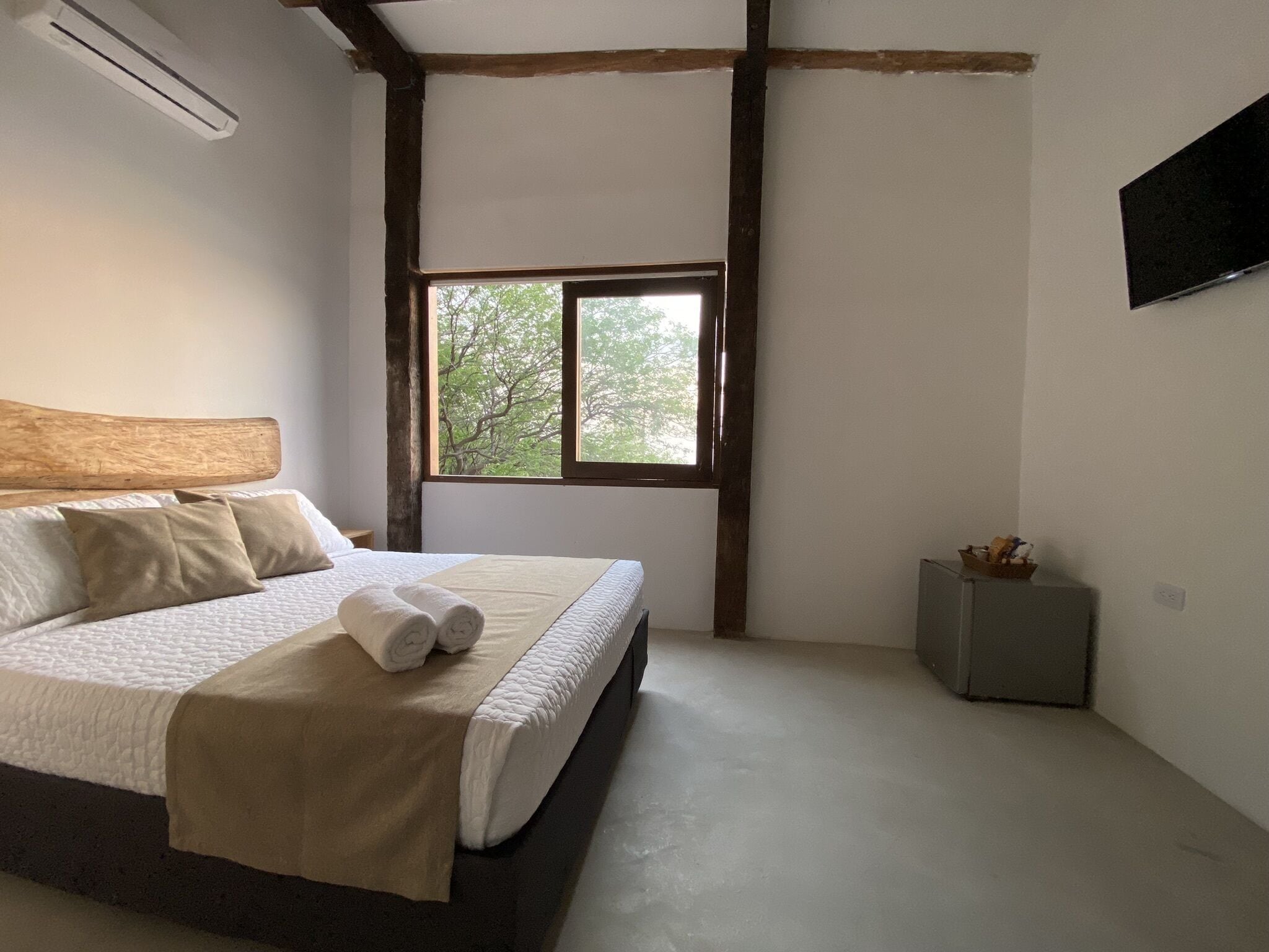 A bedroom with a large bed, a window, a small bedside table, and a flat-screen TV mounted on the wall.