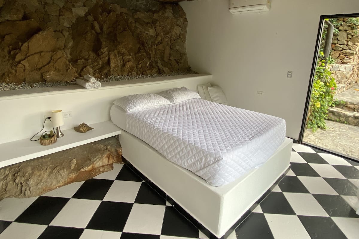 Un dormitorio con piso de cuadros, una cama con ropa de cama blanca, una pared de piedra y un estante pequeño con adornos.