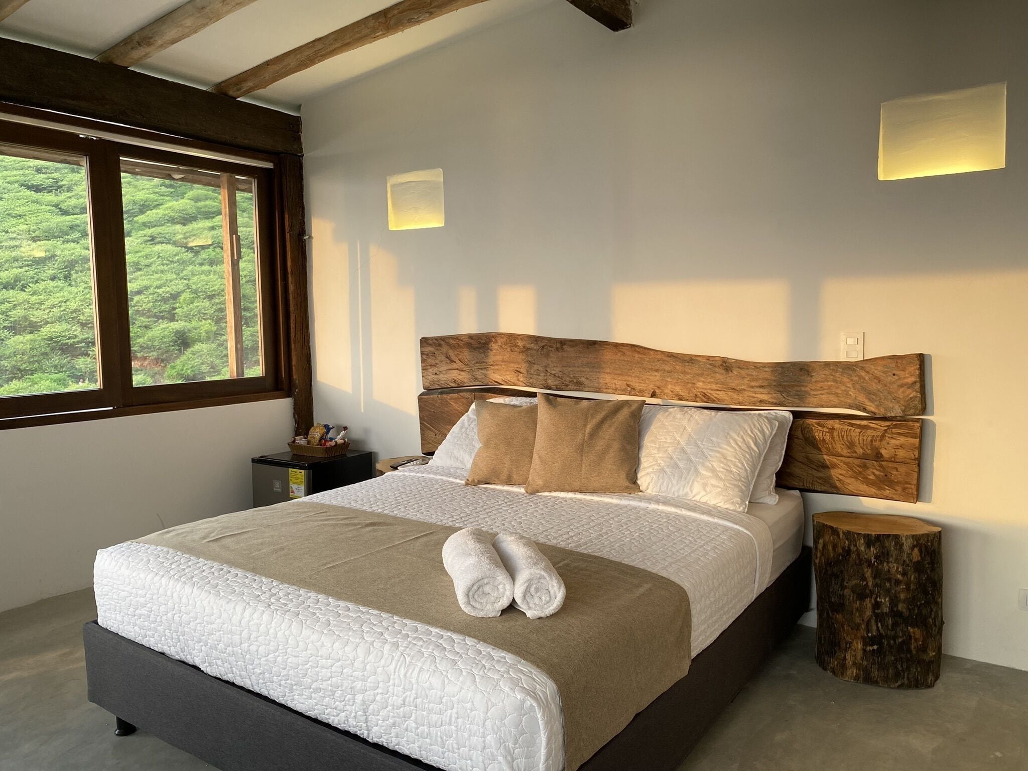 Un dormitorio con una cama grande, cabecera de madera, mesitas de noche, un taburete de madera y una ventana con vistas a la vegetación.