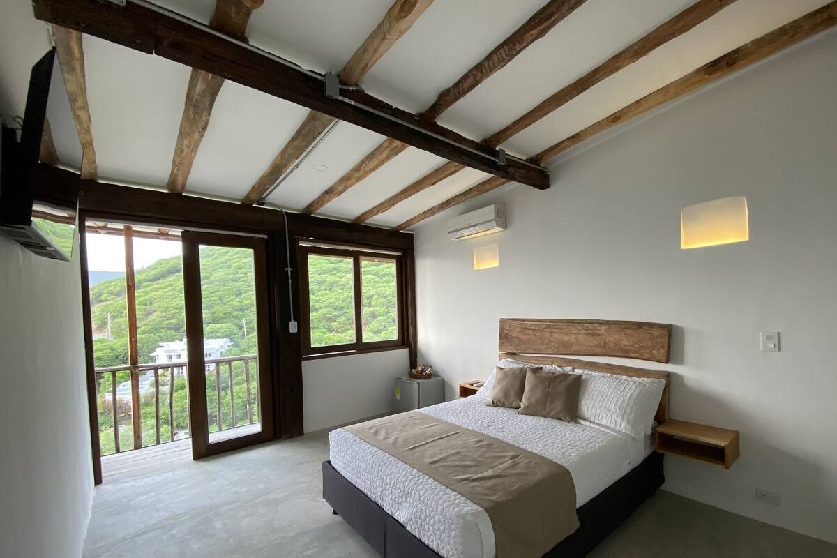 Un dormitorio con una cama grande, vigas de madera, un balcón con vistas y una mesita de noche pequeña.