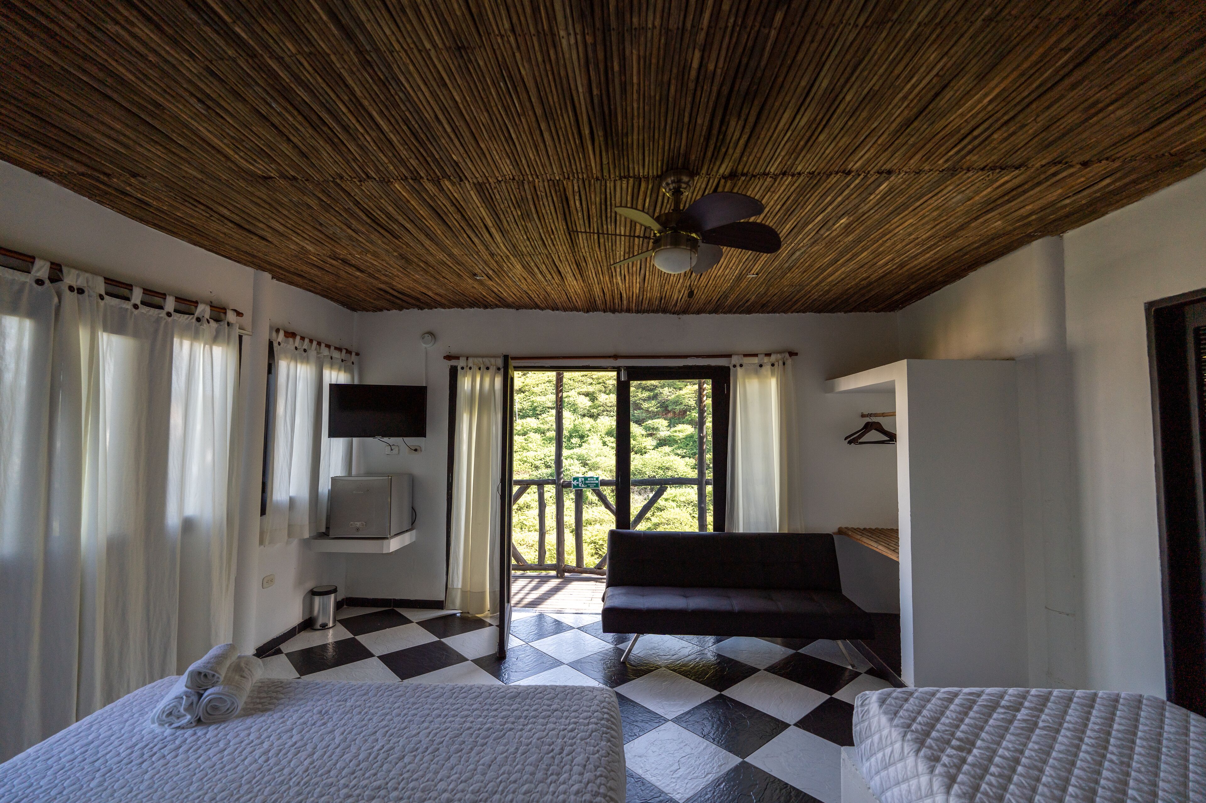 Una habitación con piso a cuadros, una cama, un sofá, un televisor y un balcón con vistas a vegetación.