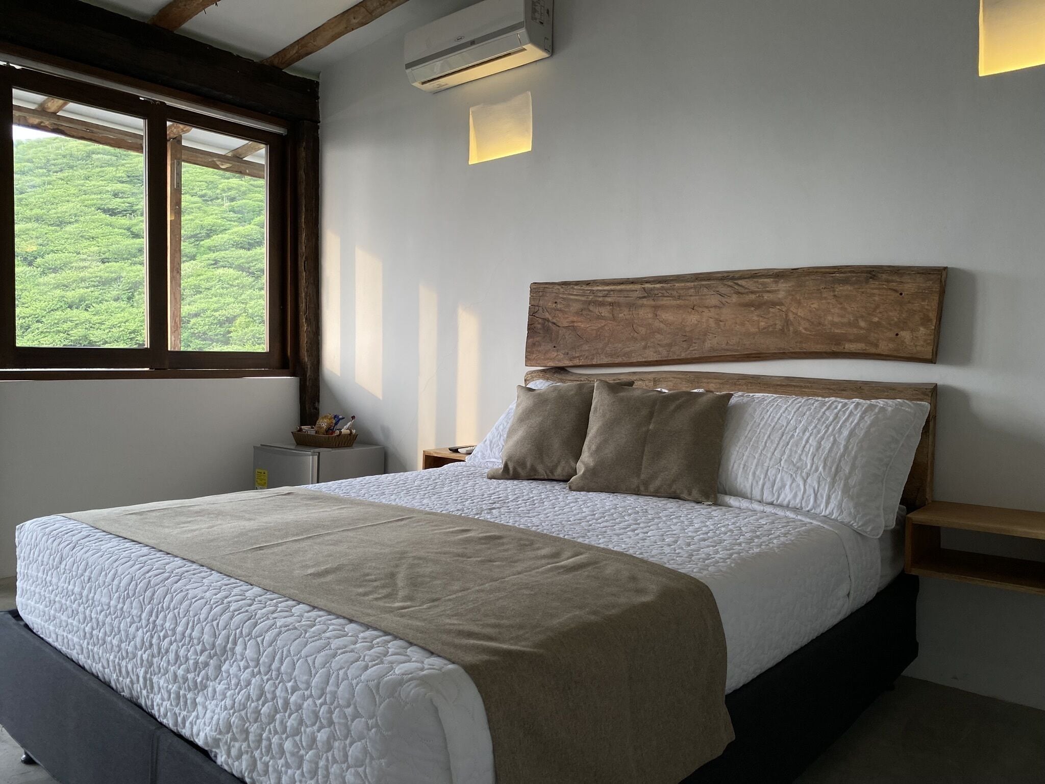 Quarto com cama grande, cabeceira de madeira, mesas de cabeceira e janela com vista para o verde.