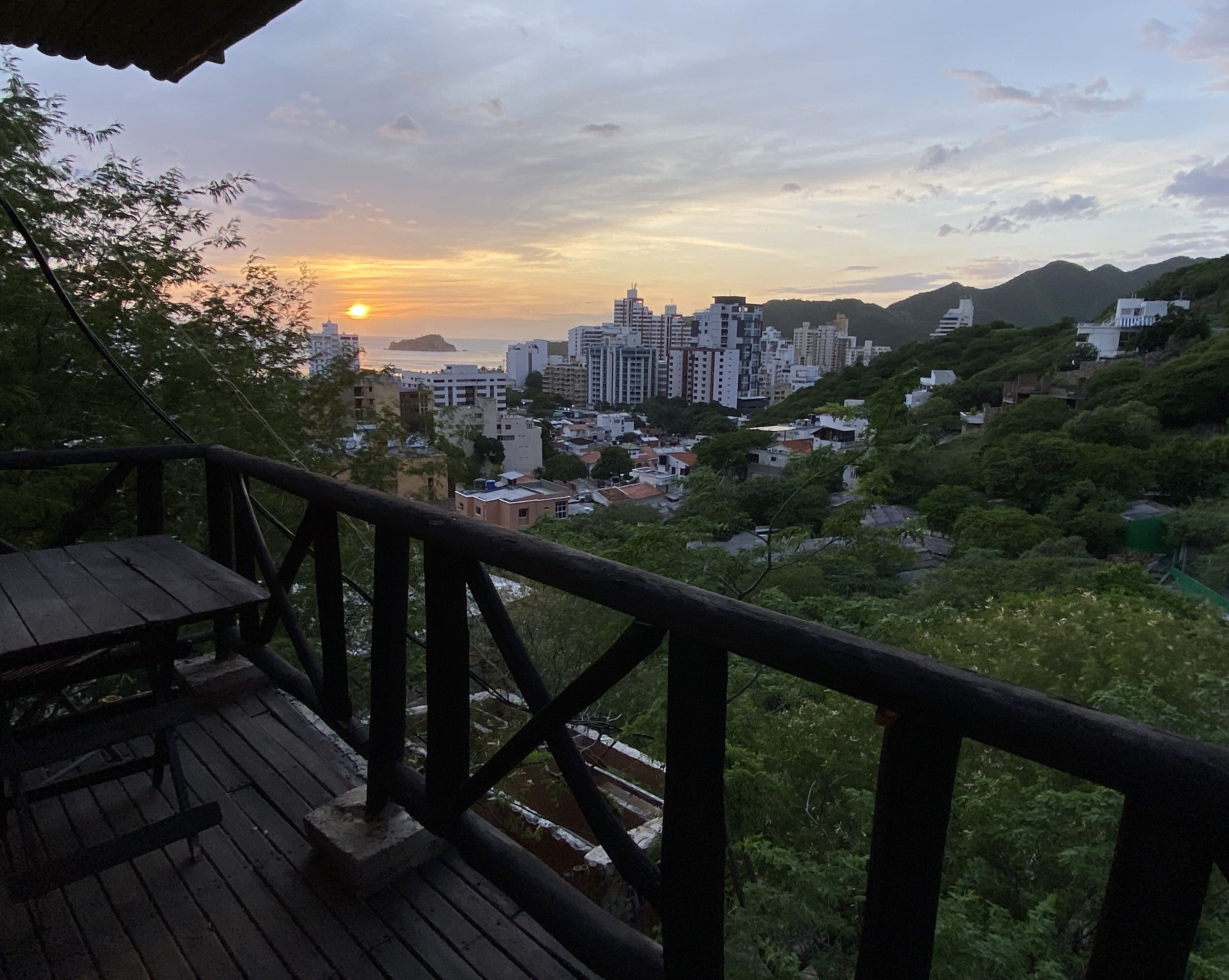 Terraza de madera con vistas a un paisaje urbano con edificios y vegetación al atardecer.