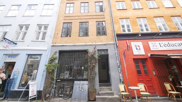 A row of buildings with a storefront on the left, a red building with a sign on the right, and a window with a bicycle in front. - LBS 3 · Copenhagen Hotel Apartments (København)