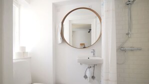 Un baño pequeño y blanco con un espejo redondo, un lavamanos y una ducha.